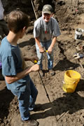 Two volunteers use probes to penetrate the surface. When the probe hits an object below, a flag is placed to mark the spot for excavation. Notice the pattern of holes created by the probe.